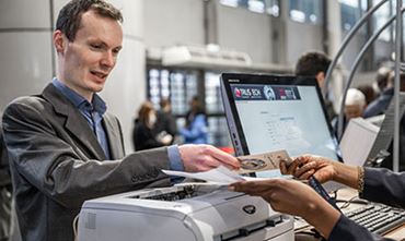 A visitor presents his pass to a trustech hostess. There is also a computer screen with the TRUSTECH registration page and a printer. 