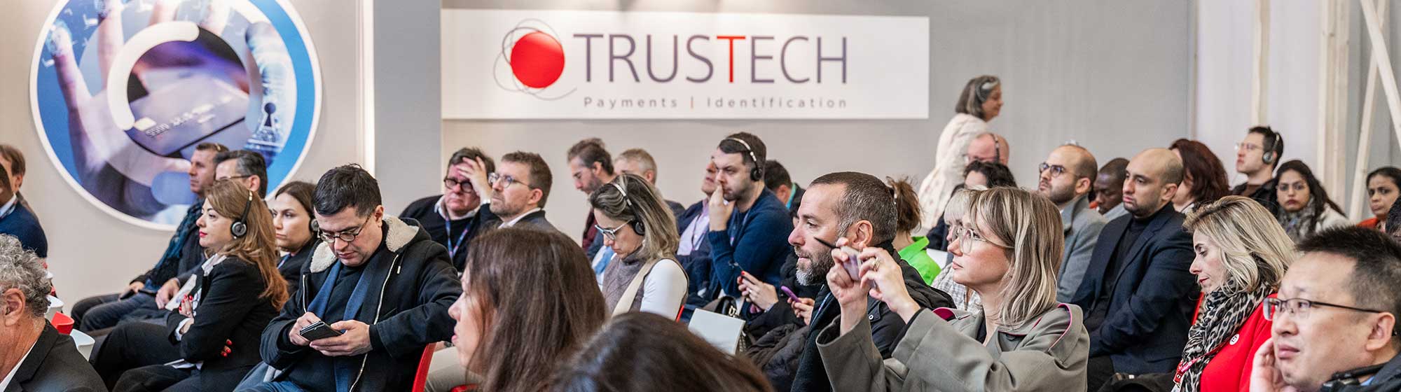 Audience seated in rows at a conference with a banner with trustech logo in the background, some attendees are looking ahead while others are using their phones.