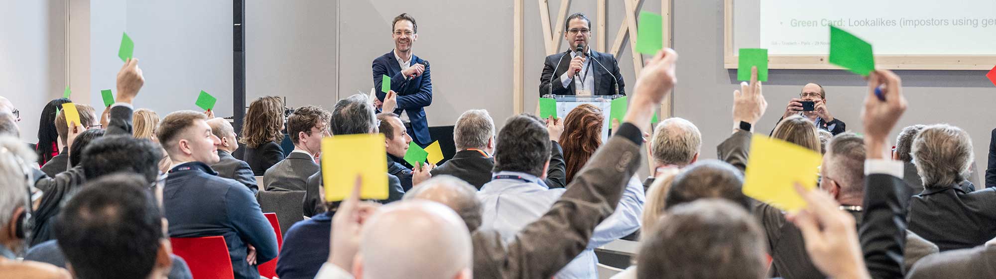 Salle de conférence où des participants lèvent des post-its verts et jaunes en réponse à une question. Deux orateurs sont présents sur scène, une présentation est affichée en retropojection