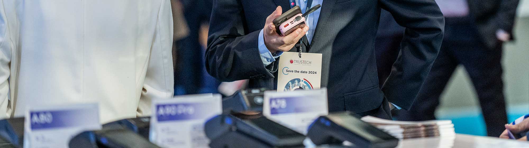 A businessman, with a Trustech badge around his neck, is looking at something off-screen, holding his phone in his hand. He is surrounded by a crowd of other visitors. Payment terminals are displayed in front of him.