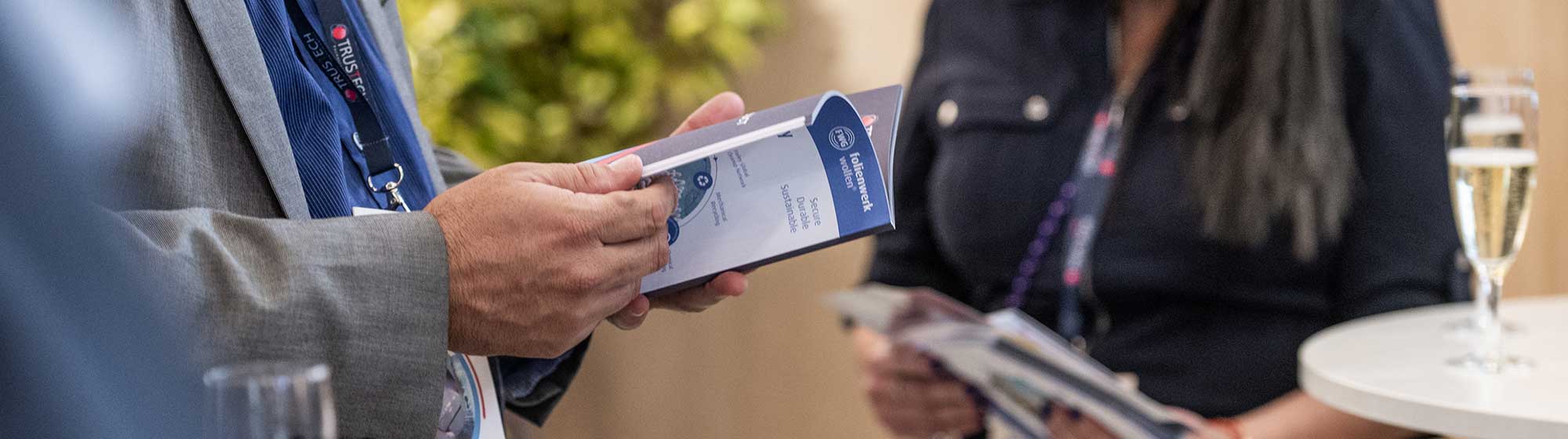 Two people at a professional event. A man in a grey suit holding a brochure titled "TRUSTECH" and a woman in a black dress sitting, holding another document. Both are wearing badges around their necks