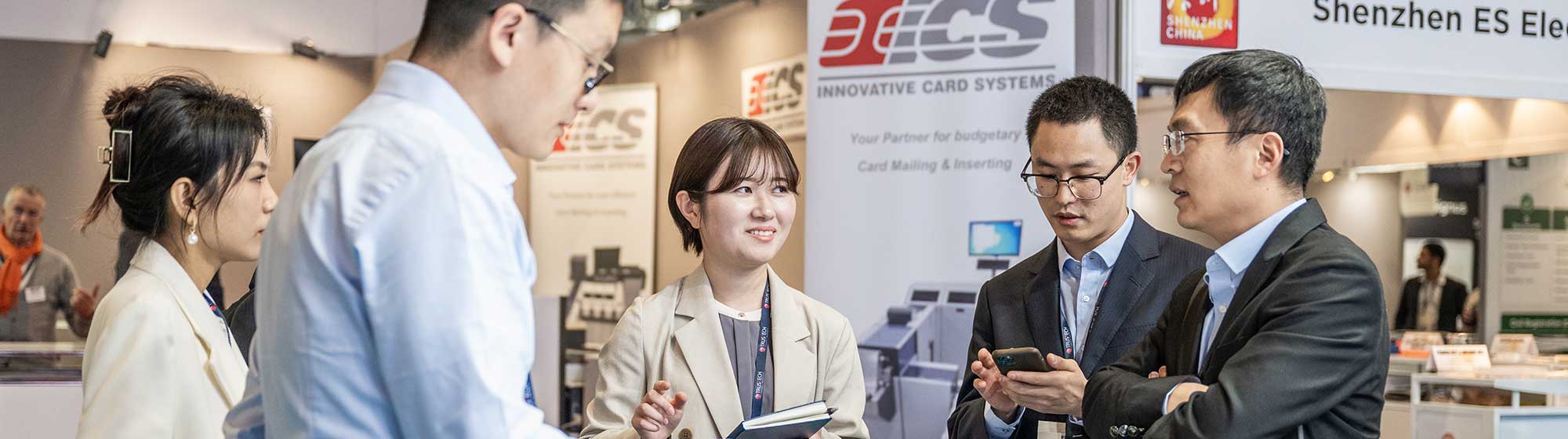 A group of professionals interacting at a booth during the TRUSTECH exhibition. They are discussing and examining various electronic devices displayed on the table. The background features banners from companies such as Innovative Card Systems and Shenzhen ES Electronics.