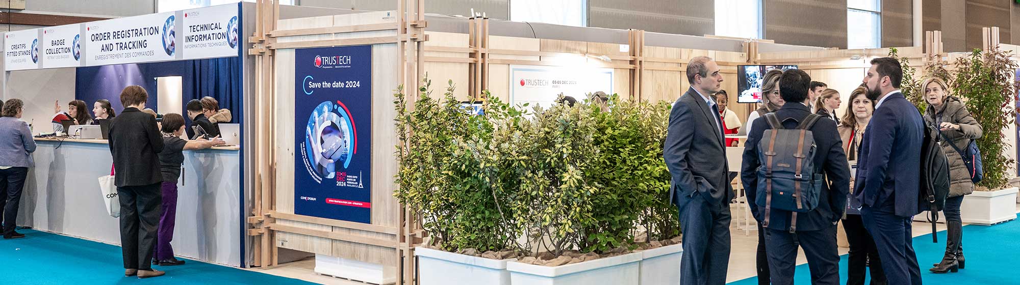 Exhibition stand at TRUSTECH 2024. Several people are interacting around the exhibitor reception stand, where services such as order registration, badge collection, and technical information are provided. Hanging signs indicate directions for various services and conference rooms. The atmosphere is formal and professional, with decor including green plants and promotional posters.