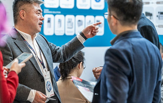 A businessman with a Trustech badge around his neck is engaging in a conversation, pointing at something off-screen. He holds a brochure in one hand and stands among other attendees, some of whom are holding their phones and reading materials. A display board with various product images and descriptions is visible in the background.
