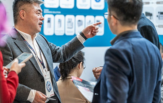 Un homme d'affaires avec un badge Trustech autour du cou est en train de converser, pointant quelque chose hors champ. Il tient une brochure dans une main et se trouve parmi d'autres participants, dont certains tiennent leurs téléphones et des documents. Un panneau d'affichage avec différentes images de produits et descriptions est visible en arrière-plan.