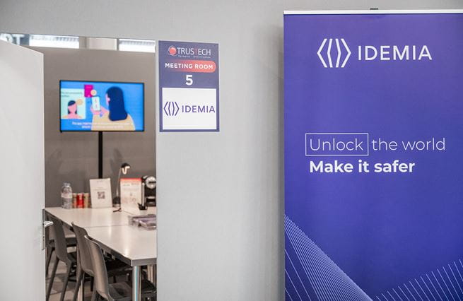 Salle de réunion sponsorisée  IDEMIA. On y voit une porte ouverte avec en arrière plan une table et un écran, et à l'extérieur un kakémono idemia sur la droite avec inscrit dessus 'unlock the world make it safer"