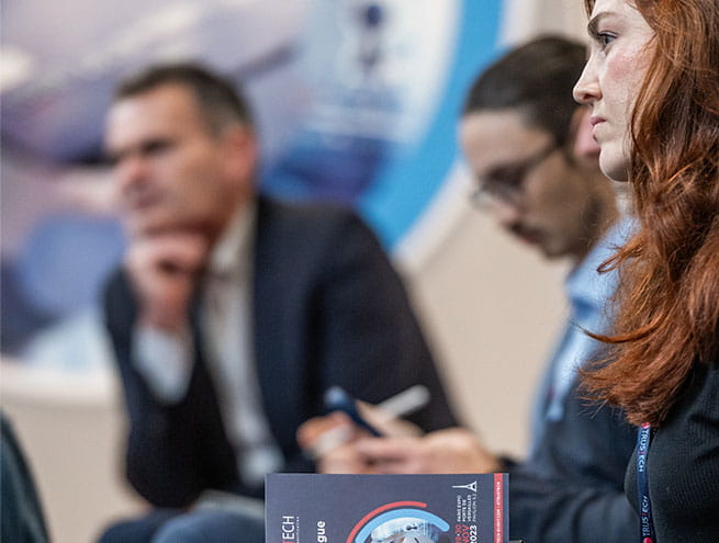 Two men and a woman assisting at a conference pitch during trustech event. The woman holds the trustech catalogue in her hands 